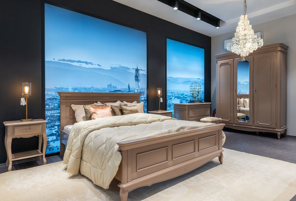 Comfortable bed with coverlet and cushions placed near nightstand and chest of drawers in spacious bedroom with crystal chandelier and screens