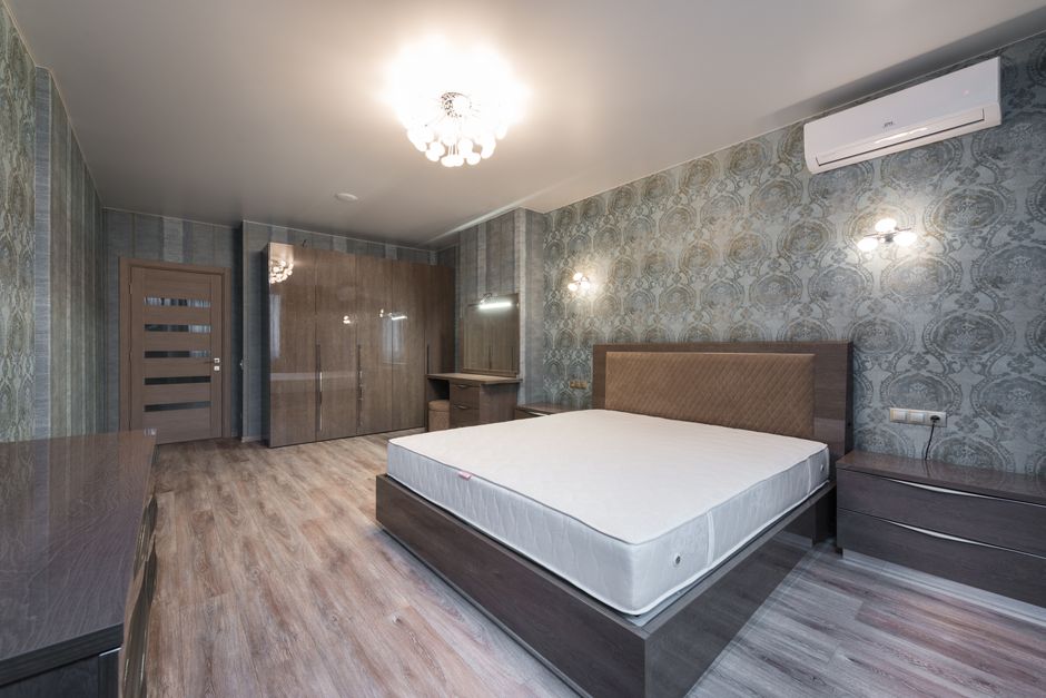 Comfortable bed with mattress placed at wall near drawer in spacious bedroom with glowing chandelier and big wardrobe near door
