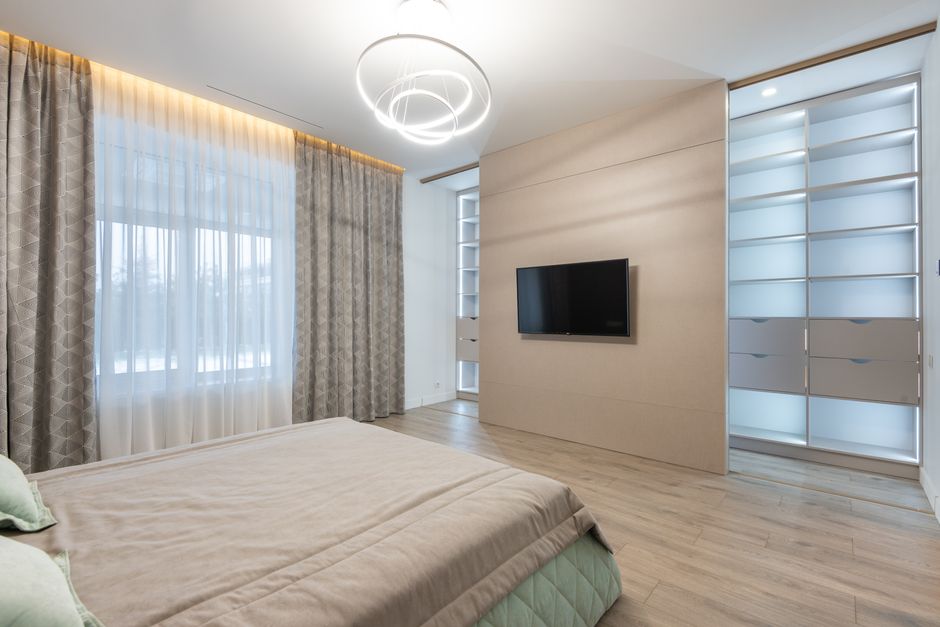 Inside bedroom interior furnished with comfortable bed covered with soft plaid against TV set placed on wall near big wardrobe with glass panels