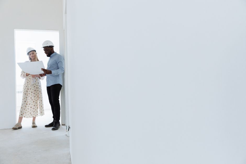 A man and woman in safety helmets examine building plans inside a minimalistic, indoor space.